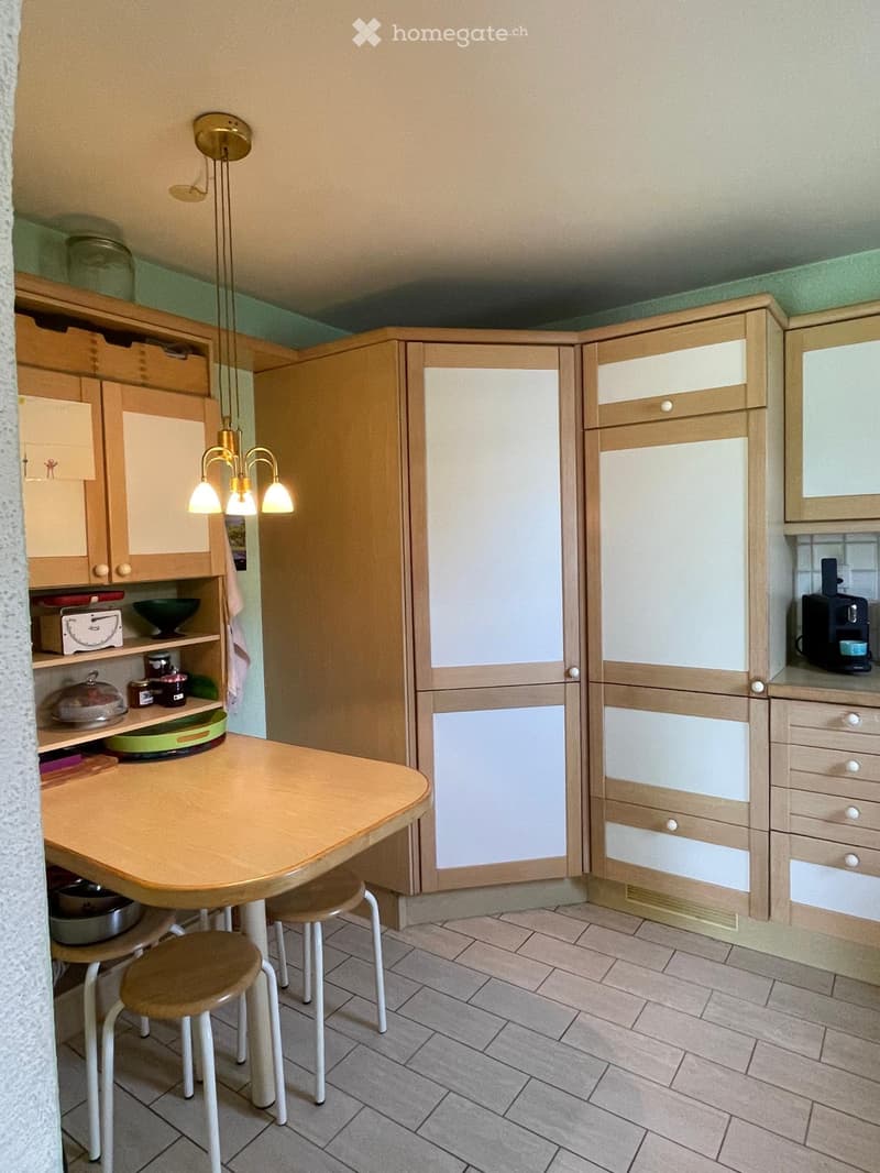 The kitchen features wooden cabinets, a central island with a round wooden table, two stools, and a refrigerator. The counter includes a coffee maker and a cup, and the shelves hold various kitchen items.