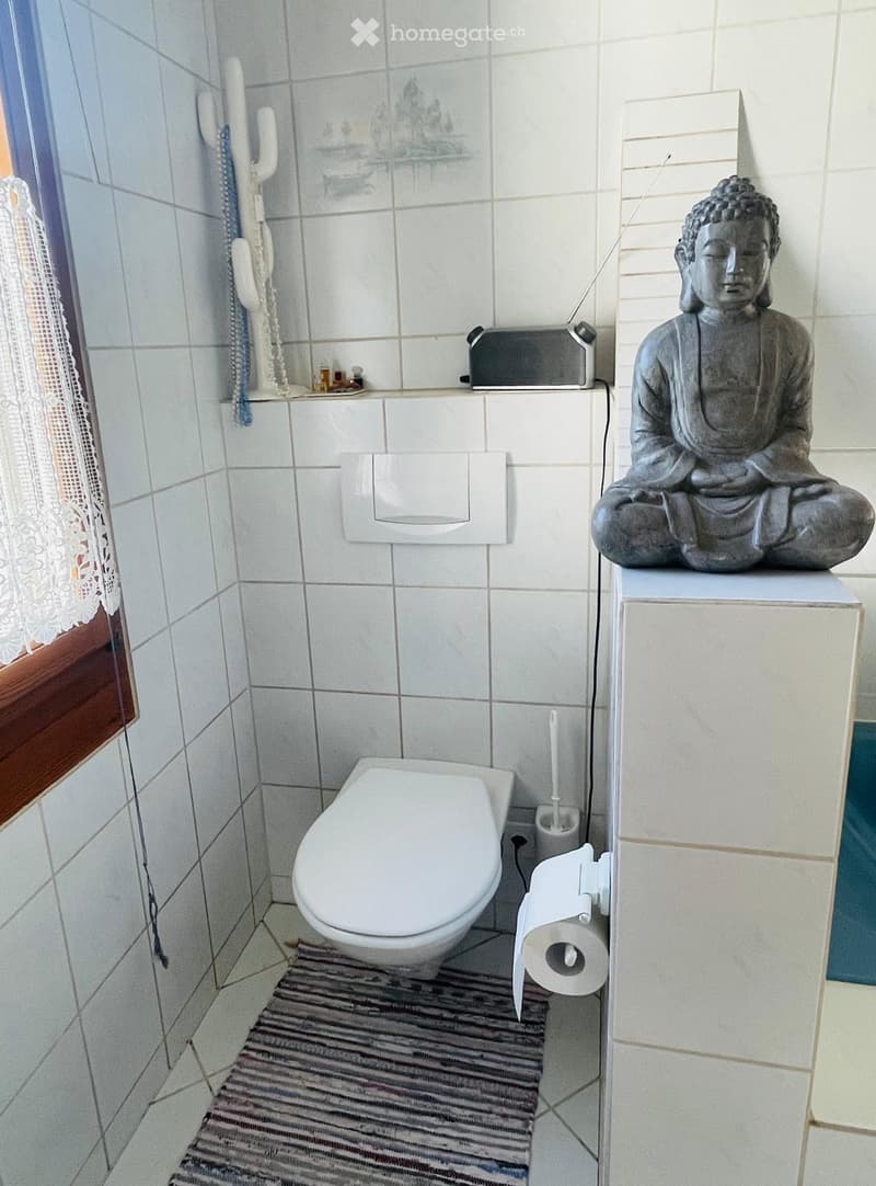 A small bathroom with a toilet, toilet paper holder, toilet brush, a small radiator, a decorative statue, and a window with a curtain.