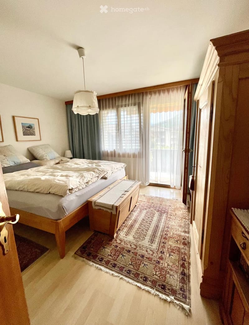 Cozy bedroom with a wooden bed, nightstand, lamp, and a large patterned rug. A sliding glass door leads to a balcony.