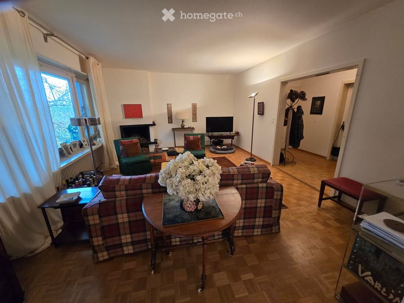 A cozy living room with a plaid-patterned sofa, coffee table with a floral centerpiece, armchairs, fireplace, and a television on a stand. The room features wooden flooring and large windows with curtains.