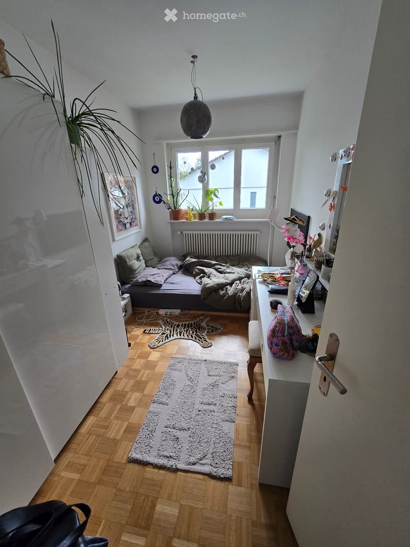 Cozy bedroom with wooden flooring, a sofa bed, a small table, a chair, a framed picture, a rug, and a hanging lamp.