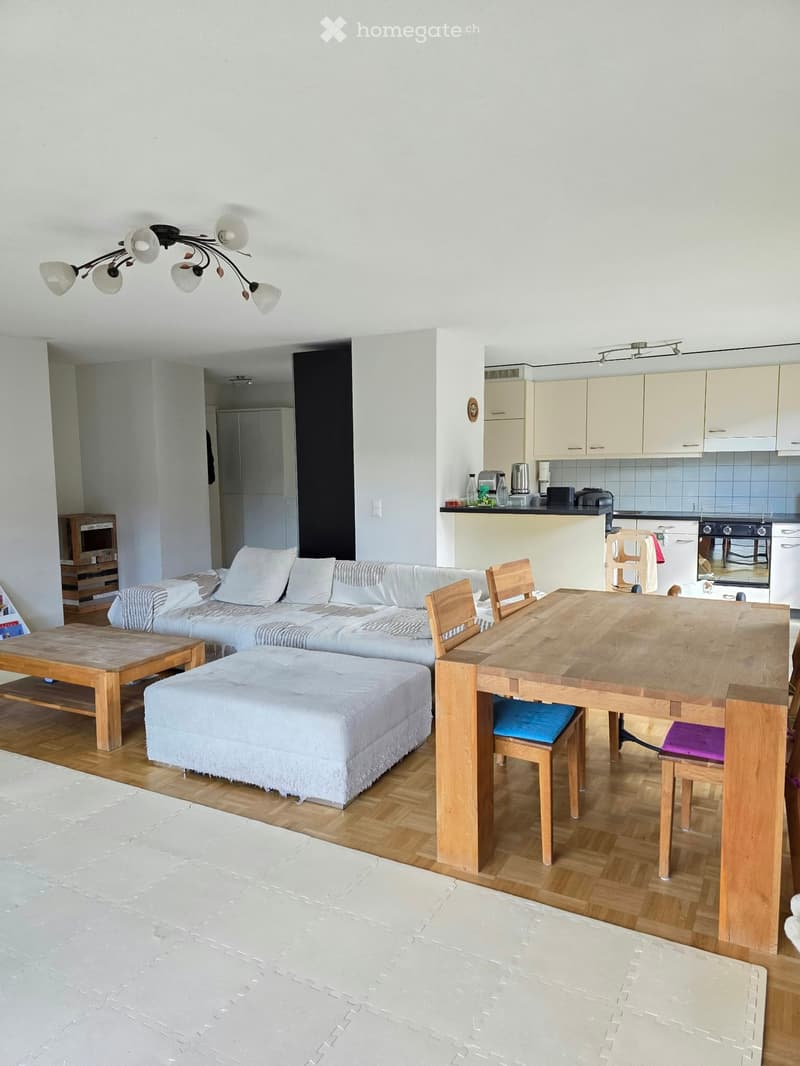 Bright, open-plan living and dining area with wooden flooring, white walls, and modern furniture including a sofa, ottoman, coffee table, dining table with chairs, and a small kitchen area with cabinets and appliances.