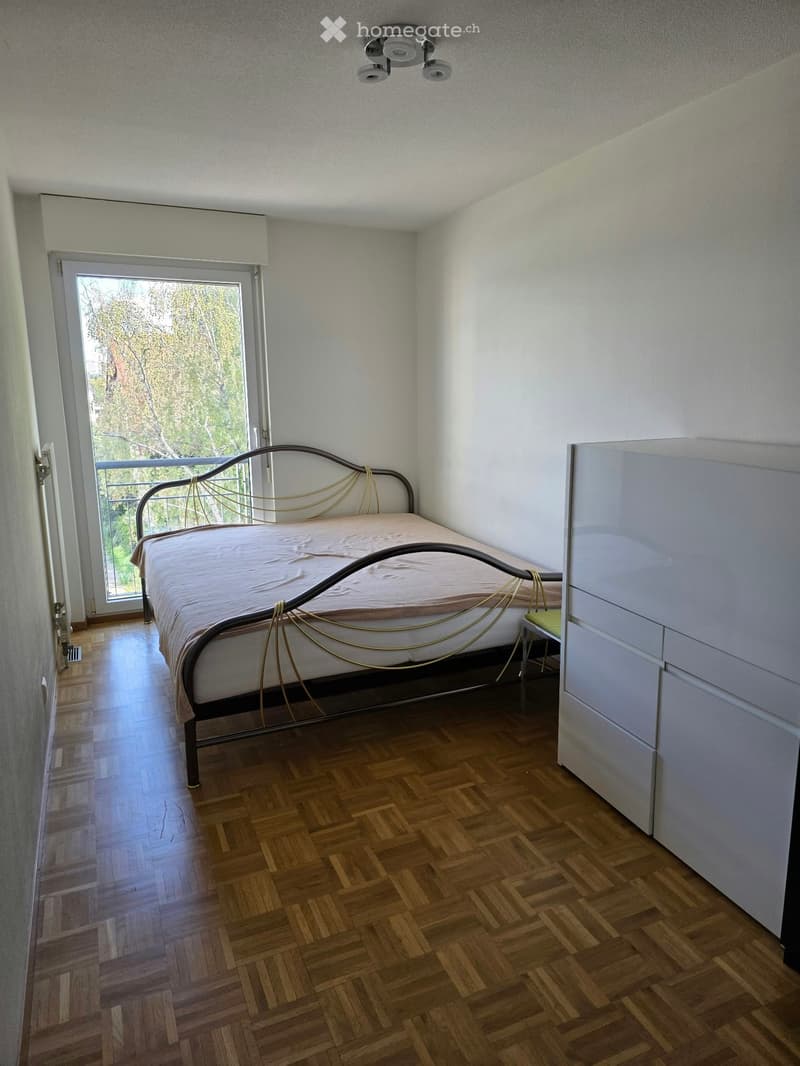 Bedroom with wooden herringbone flooring, a metal-framed bed with a beige bedspread, and a white wardrobe. A large window with blinds offers a view of greenery outside.