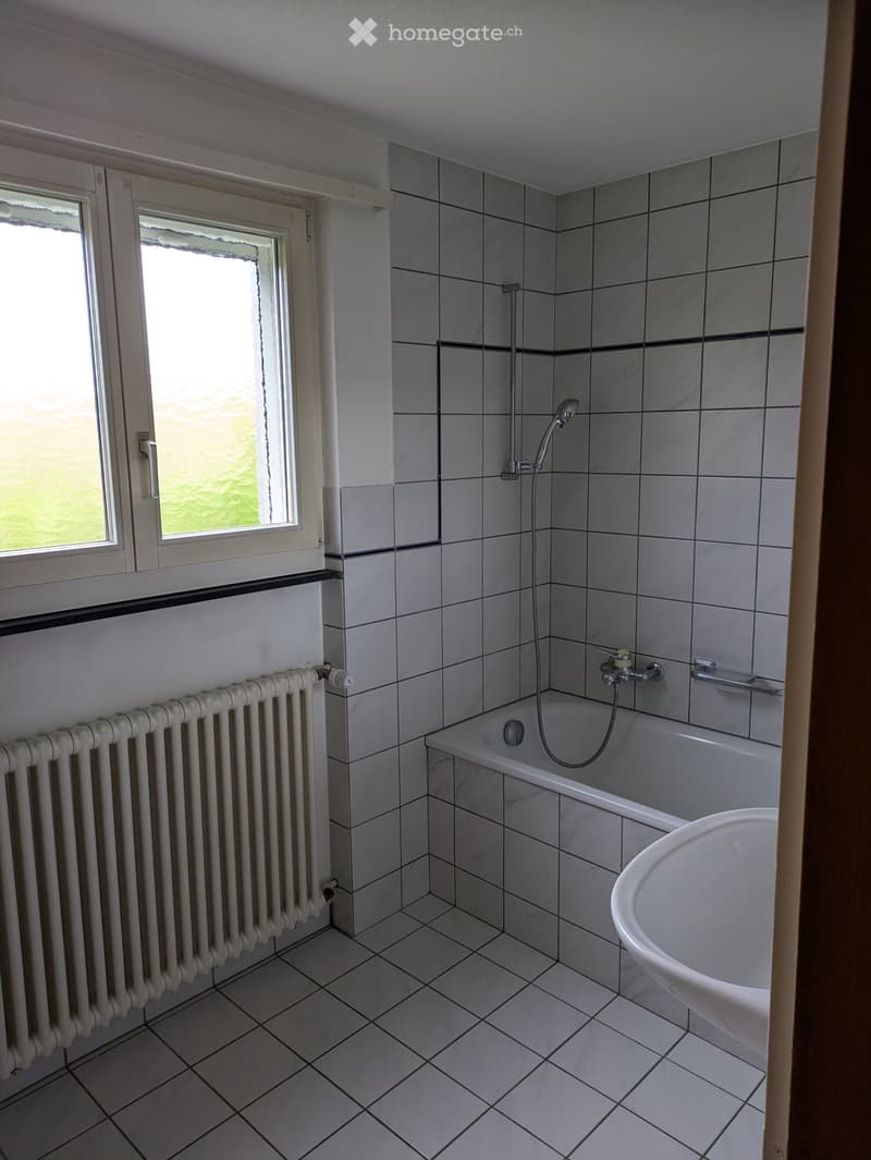 Bathroom with tiled walls and floor, bathtub with showerhead, sink, radiator, and window providing natural light.