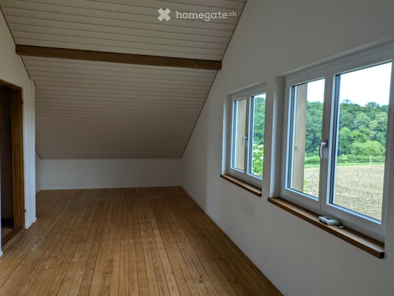 An empty attic room with wooden flooring, white walls, and large windows offering a view of greenery outside. The ceiling has exposed wooden beams, and there is a small wooden shelf beneath one of the windows.