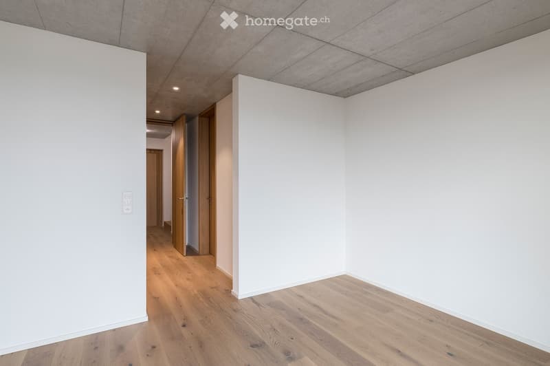 Empty room with white walls, wooden flooring, and recessed lighting. Features a doorway leading to another room with wooden doors.