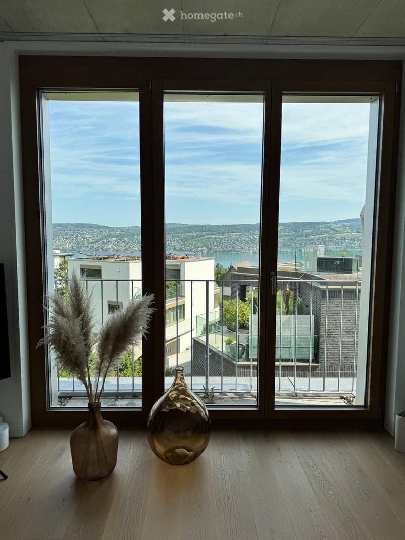 Bright room with large windows offering a scenic view of a lake and surrounding landscape. Two decorative vases are placed on the wooden floor.