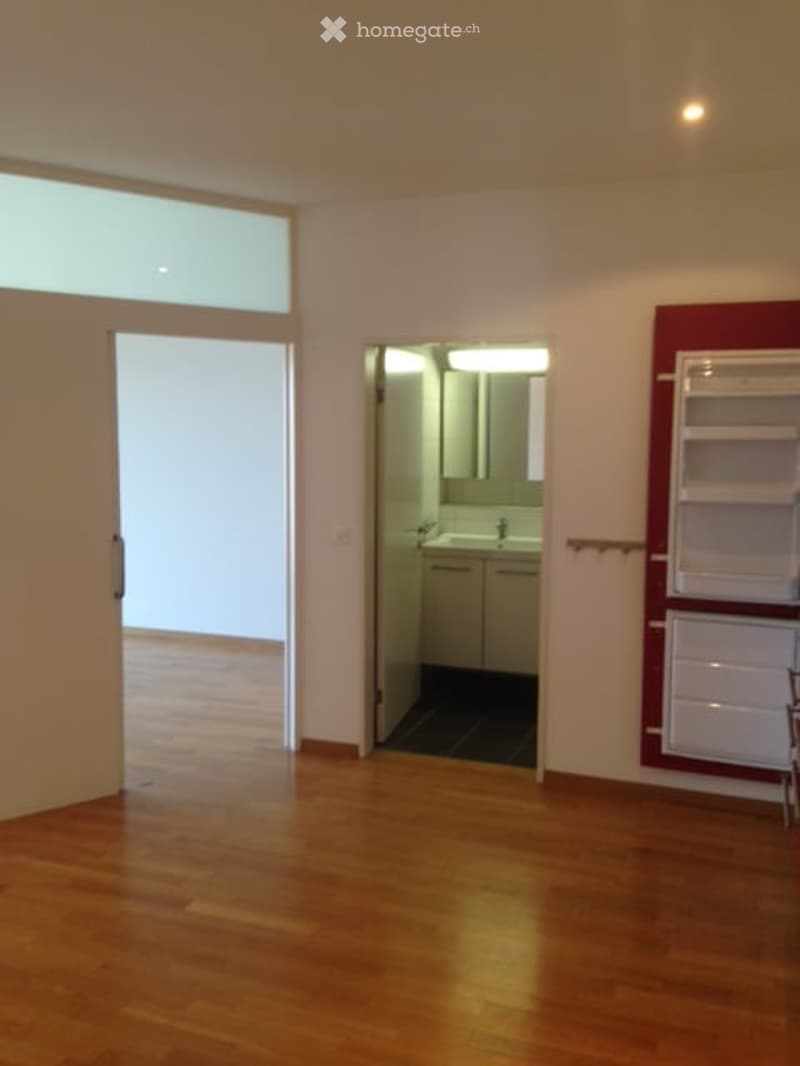 Empty room with wooden flooring, large window, open doorway leading to a small bathroom with white cabinets and a sink.
