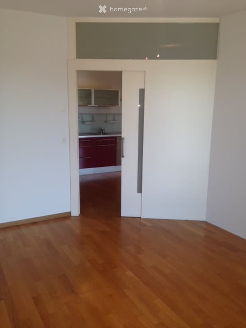 Empty room with wooden flooring and white walls, featuring a sliding door leading to a kitchen area with red cabinets and stainless steel appliances.