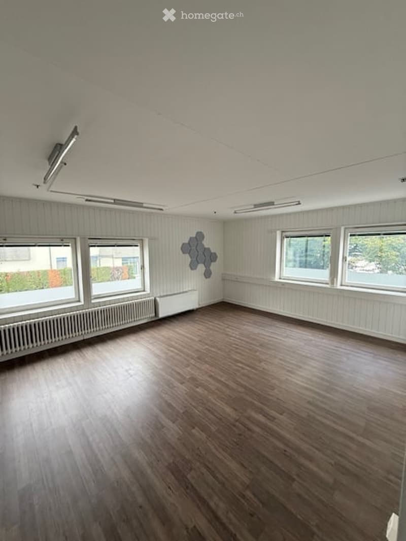 Empty room with wooden flooring, white paneled walls, and large windows. A decorative wall piece is visible on one wall.