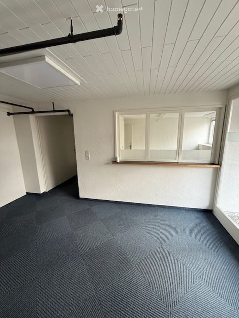 Empty room with white walls, blue carpet, large windows, and a wooden shelf. No furniture or equipment present.
