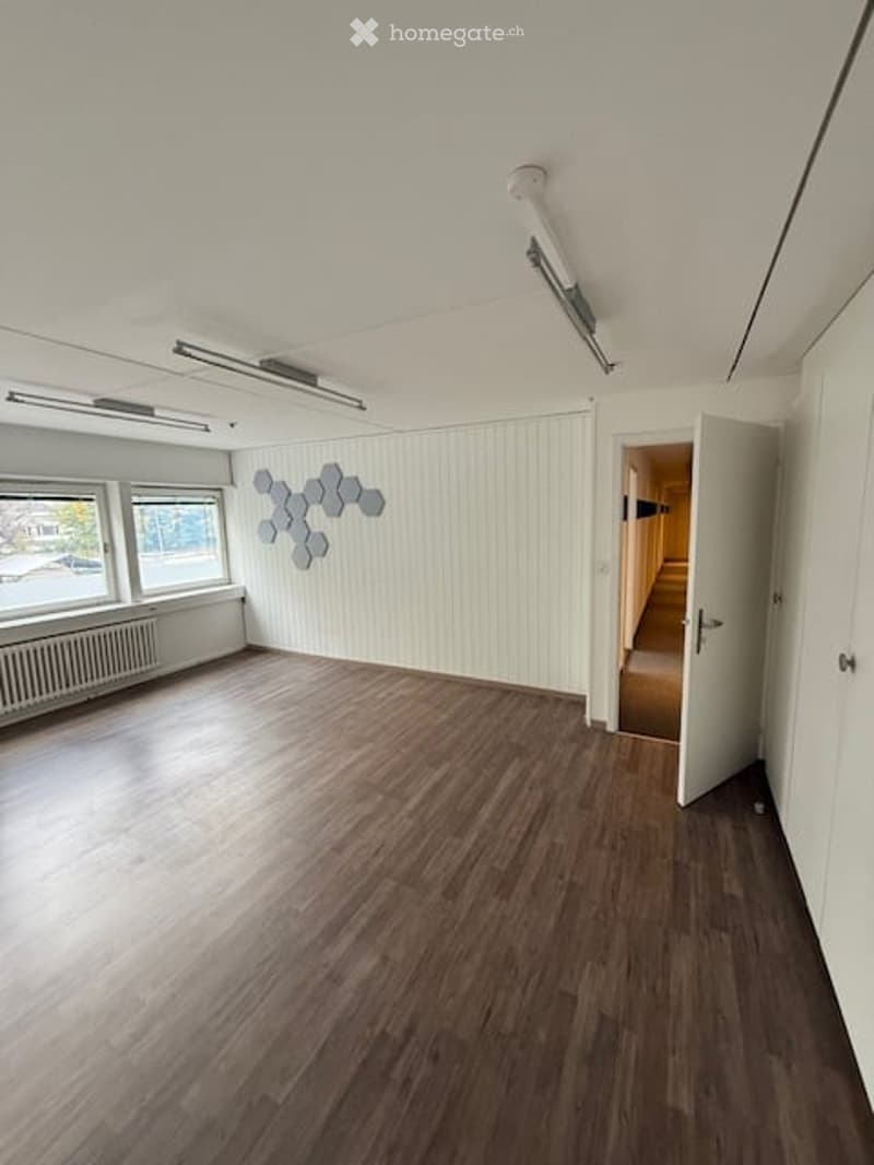 Empty room with wooden flooring, white paneled walls, large windows, and a modern geometric wall decoration. No furniture or equipment present.