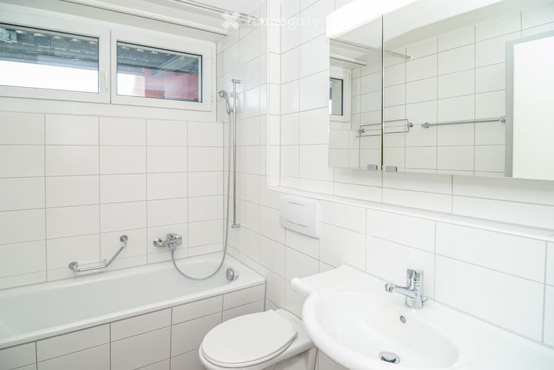 Modern bathroom with white tiles, bathtub, toilet, sink, mirror, and showerhead.