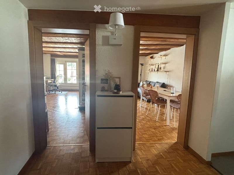 A well-lit hallway with wooden flooring and exposed wooden beams on the ceiling. The hallway leads to a dining area with a table and chairs, and a living area with a sofa and shelves. A small refrigerator is positioned against the wall in the hallway.