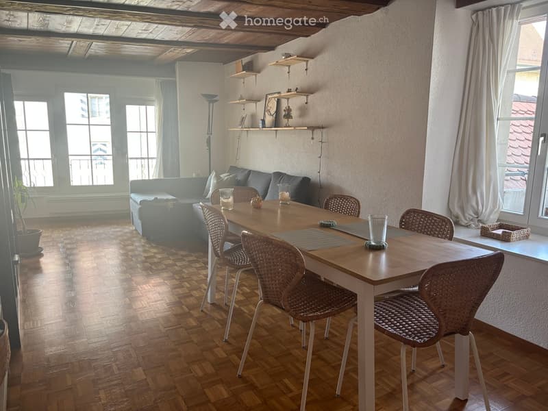 Bright living room with wooden flooring, large windows, a grey sofa, a dining table with four wicker chairs, and decorative shelves.