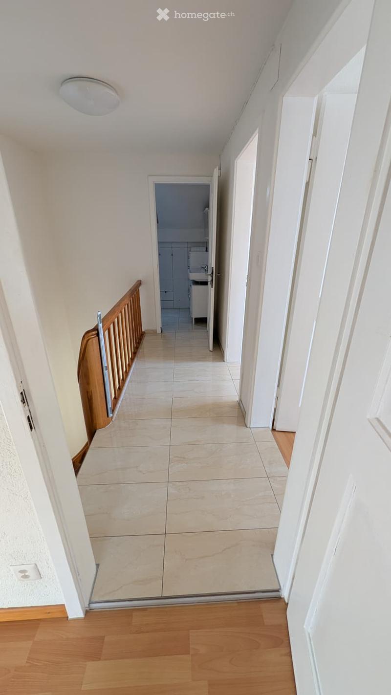 A clean, well-lit hallway with light-colored tiled flooring, white walls, and wooden railings. An open door leads to a bathroom with white cabinets and a sink.