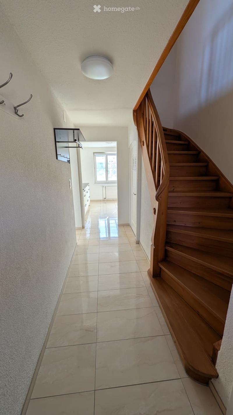 A narrow hallway with tiled flooring, a wooden staircase on the right, and a small mirror on the left wall. The ceiling has a round light fixture, and a doorway leads to another room with white cabinets and a window.