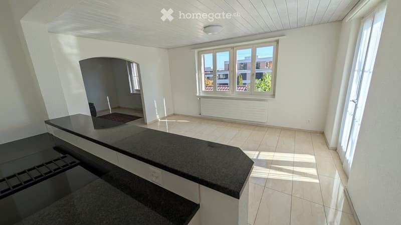 Empty room with white walls, tiled floor, large windows, and a black countertop in the foreground.