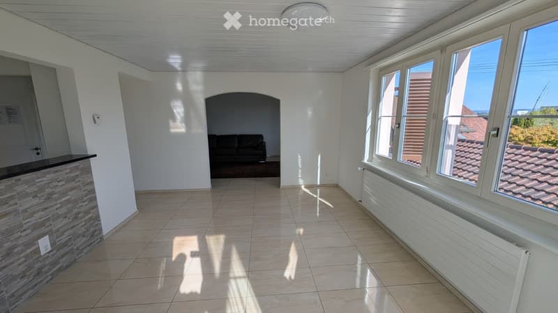 Bright living room with large windows, tiled floor, white walls, and a dark-colored sofa visible in the background.