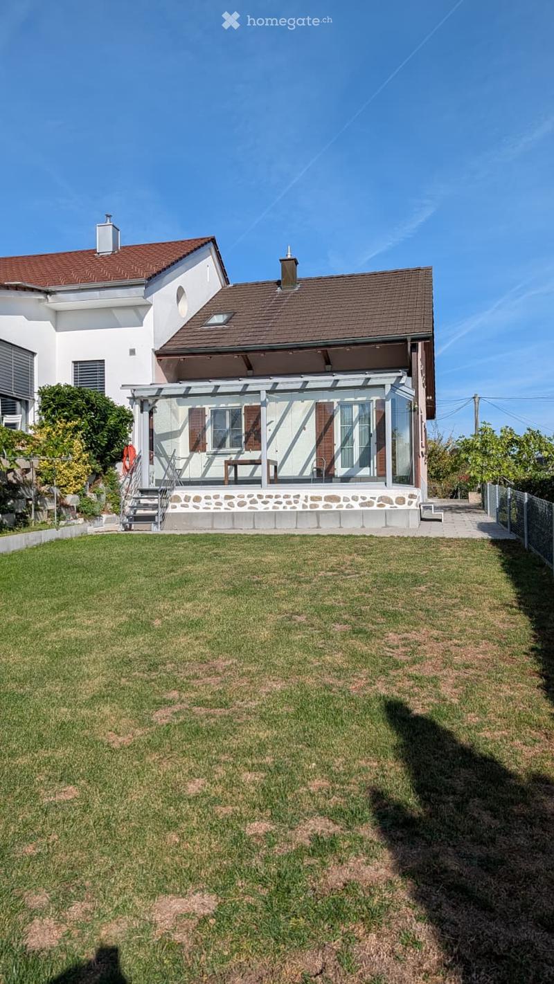 Two-story house with a red-tiled roof, white walls, and a covered patio area with a table and chairs. The house is surrounded by a well-maintained lawn and trees.