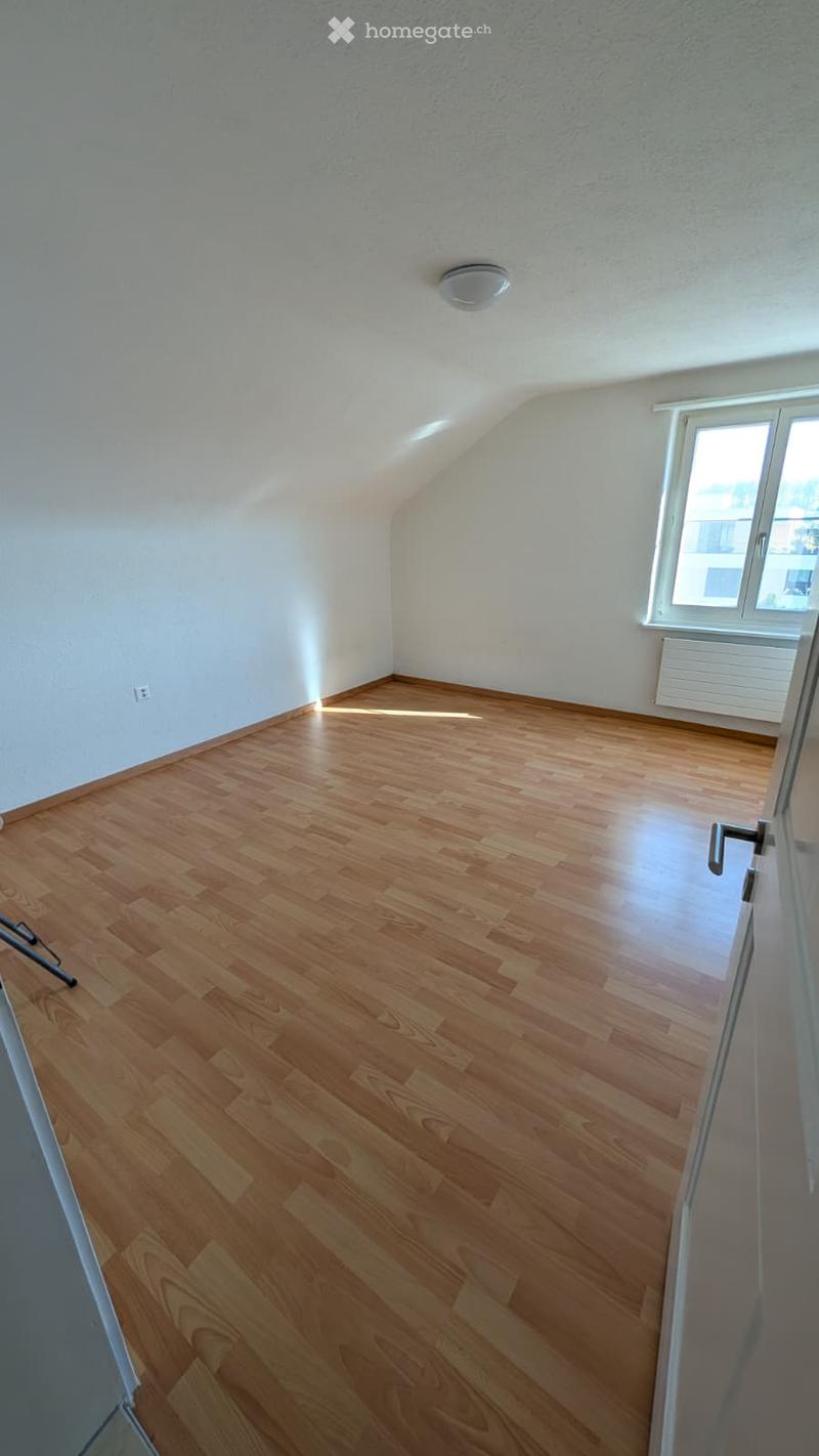 Empty room with wooden flooring, white walls, and a large window letting in natural light. The room has a slanted ceiling typical of an attic space.
