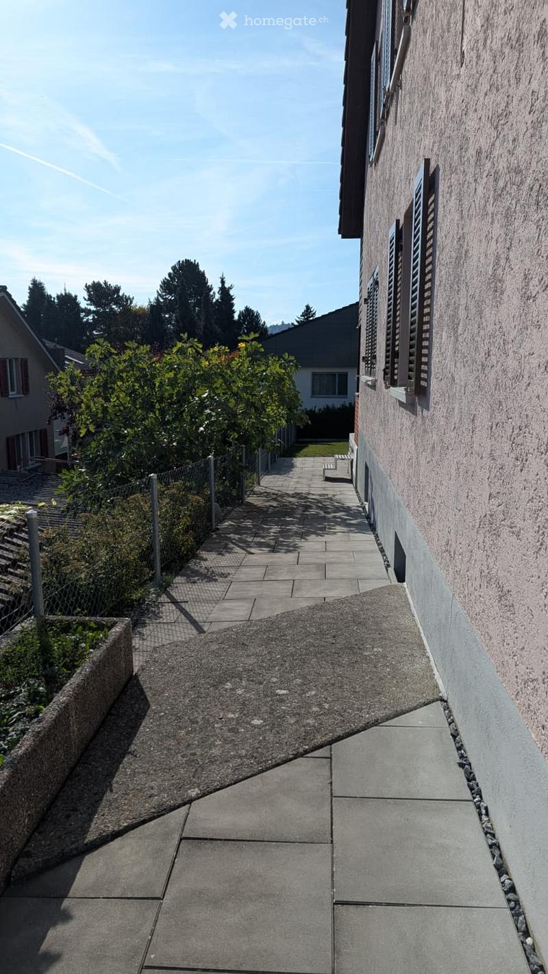 A narrow, paved pathway leads to a house with a stone facade and shutters. The pathway is bordered by a low fence and greenery, with a small bench visible near the house.