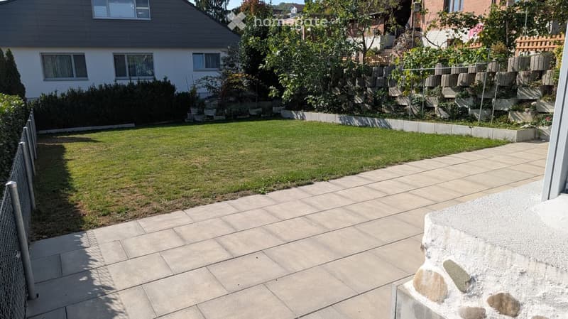 Well-maintained garden with a neatly trimmed lawn, stone pathway, and surrounding greenery, including shrubs and trees. The garden is bordered by a low stone wall and a metal fence.