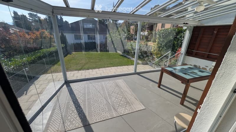Bright, enclosed patio with glass walls and roof, featuring a small table and chair, a patterned rug, and a view of a well-maintained garden.