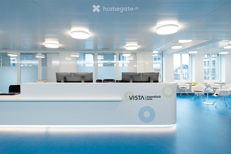Modern office space with a reception desk, multiple computer screens, chairs, and tables arranged around the area.