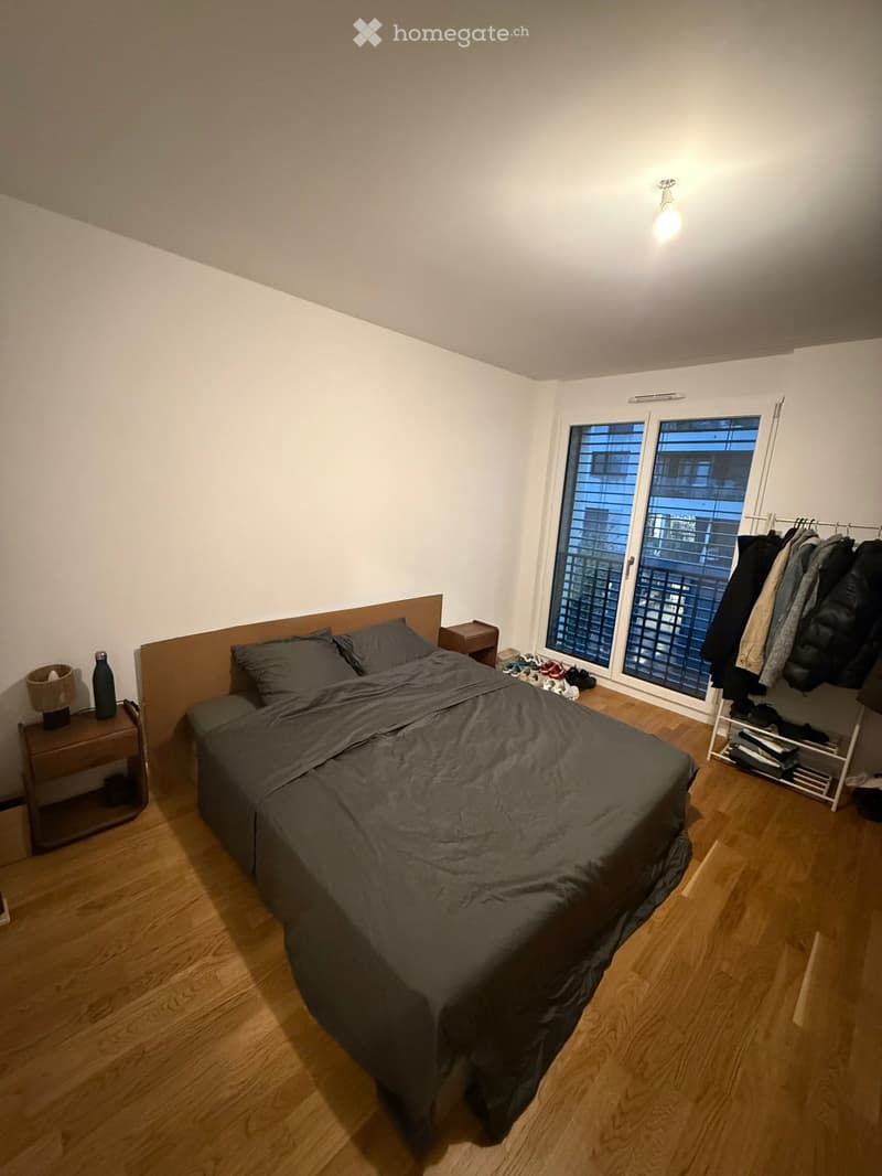 A modern bedroom with a wooden floor, a large bed with grey bedding, a nightstand with a lamp and bottle, a wardrobe with hanging clothes, and a pair of shoes on the floor near the window.