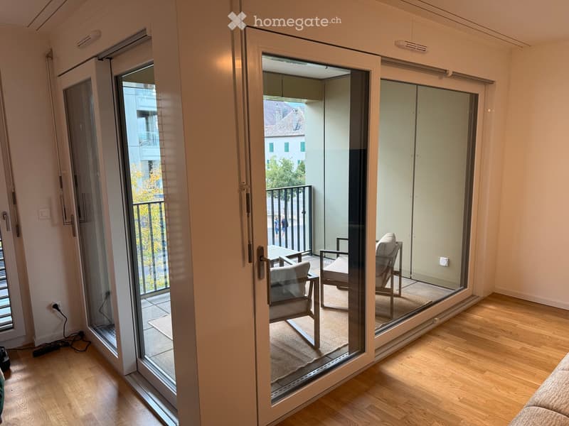 A modern living room with wooden flooring, large glass sliding doors leading to a balcony, and a few pieces of furniture including a chair and a small table.