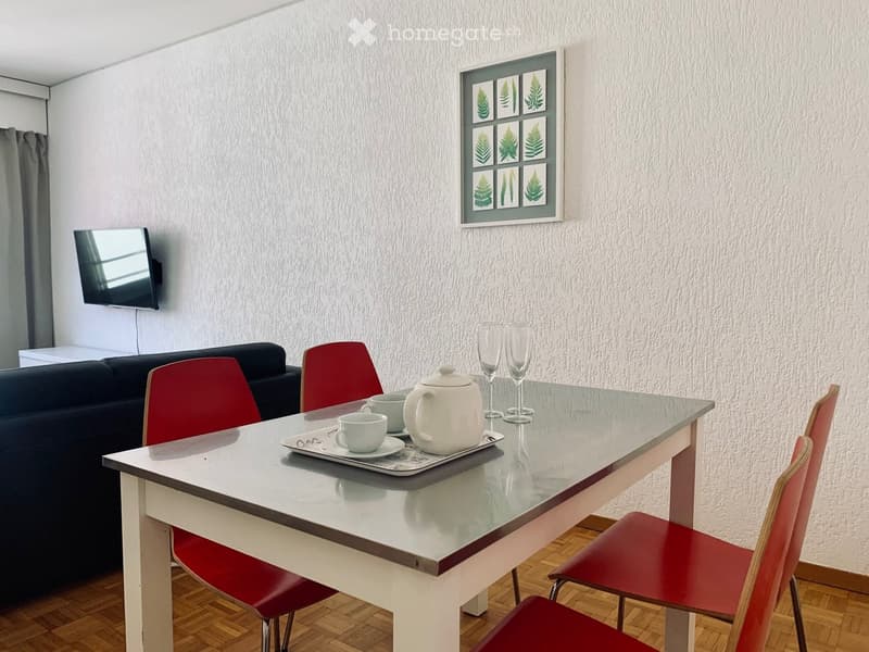 A modern living room with a glass table, red chairs, a black sofa, a teapot, cups, and a TV screen on the wall.