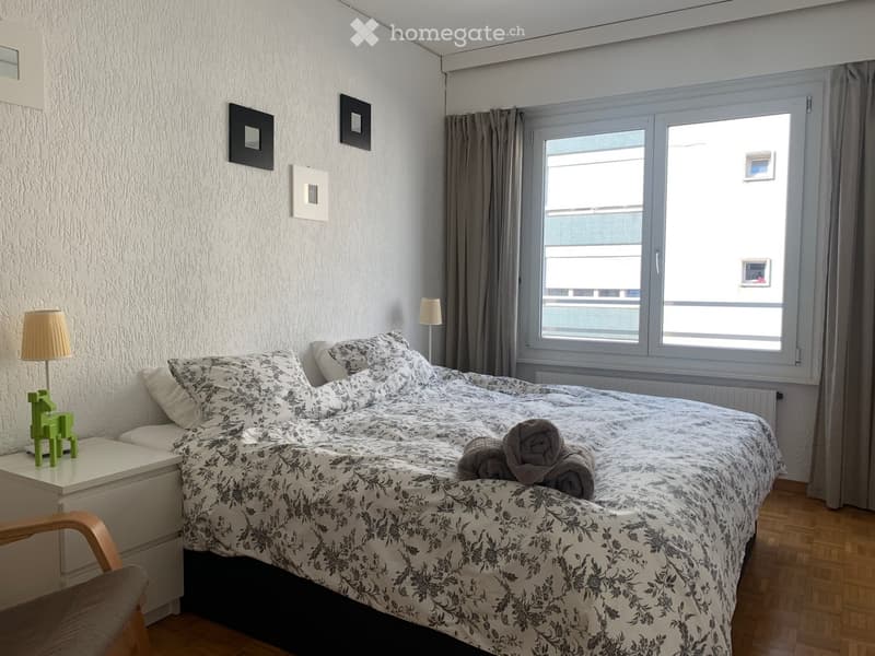 Cozy bedroom with a double bed, nightstand, lamp, and a small decorative green item on the nightstand. The room features wooden flooring and large windows with curtains.