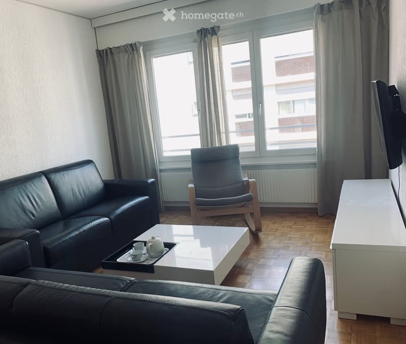 Modern living room with a black leather sofa, a white coffee table, a single armchair, and a tray with a teapot and cups on the coffee table. The room has large windows with curtains and wooden flooring.