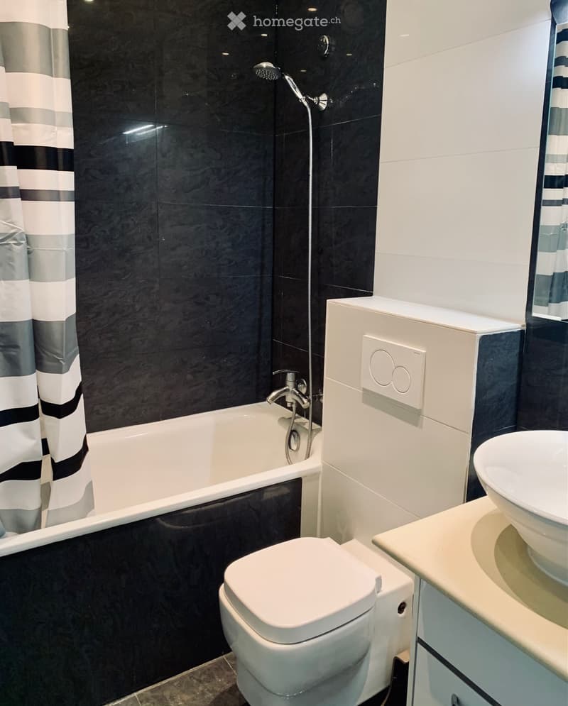 Modern bathroom with black tiled walls, white bathtub, toilet, sink, and showerhead.