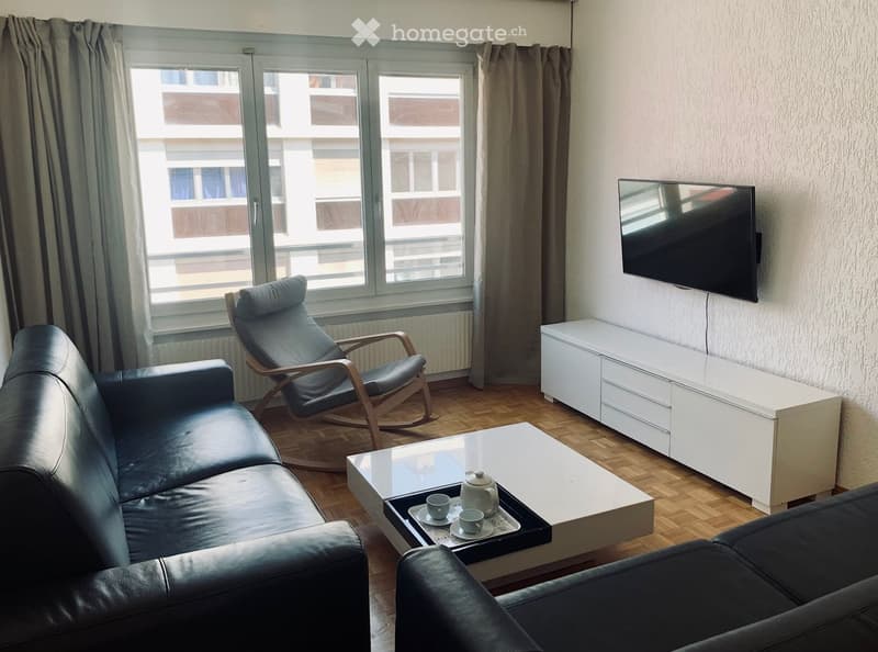 Modern living room with a dark leather sofa, white coffee table, a rocking chair, a TV mounted on the wall, and a white TV stand.