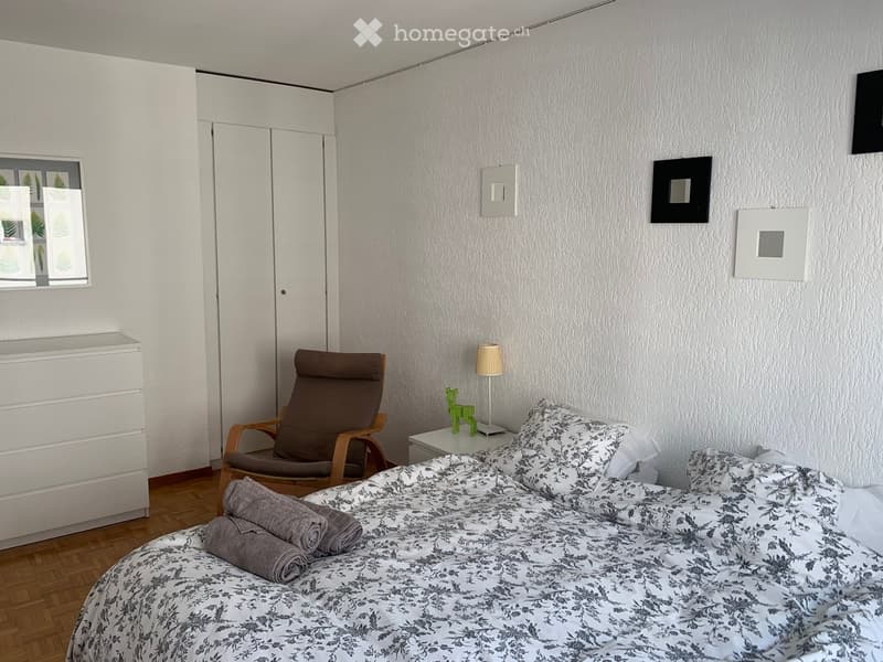 Modern bedroom with a double bed, patterned bedding, a wooden chair, a small table with a lamp, and a folded towel on the bed.