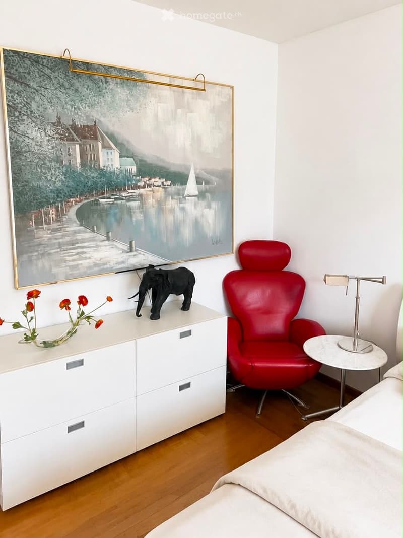 Modern bedroom with a large bed, white dresser, red armchair, small round table, vase with flowers, elephant figurine, and a large picture frame on the wall.