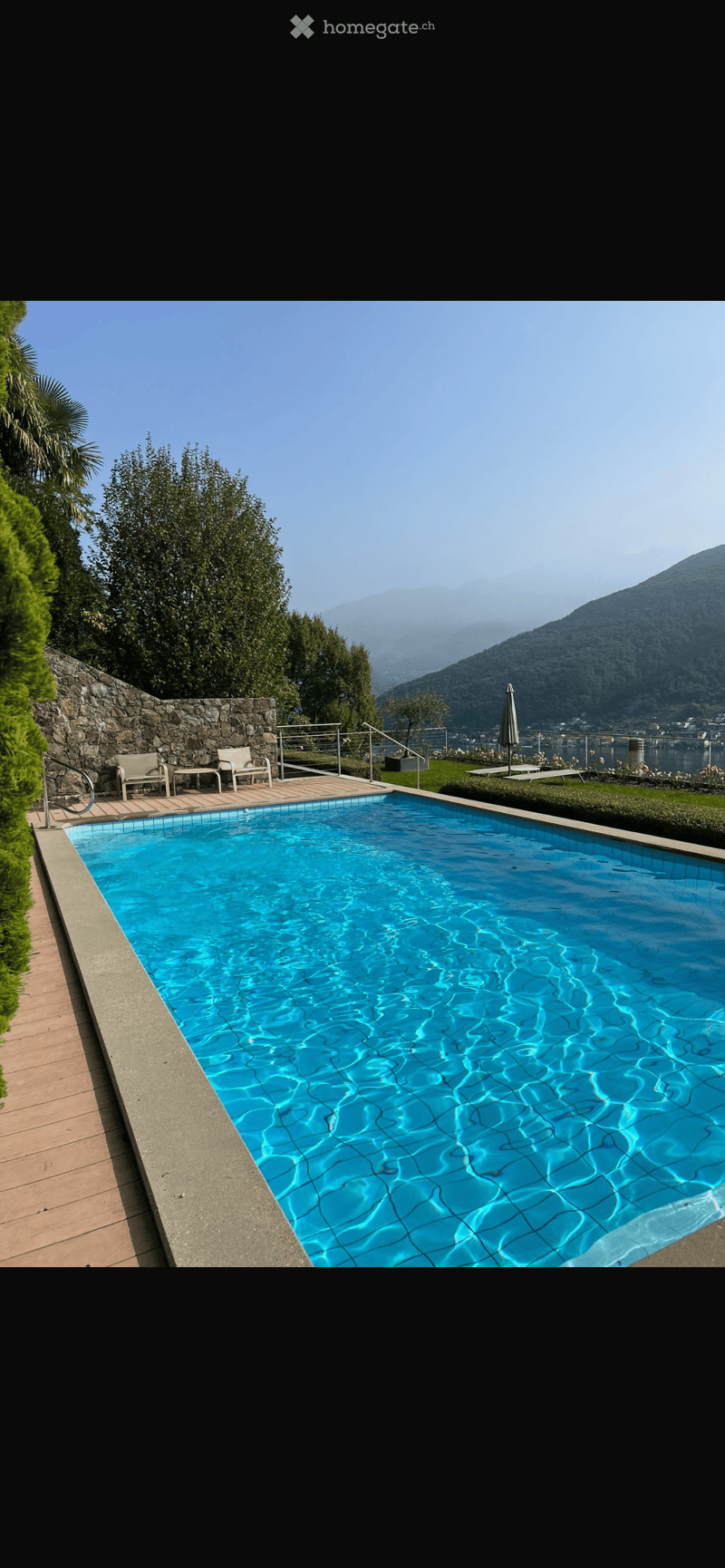 A rectangular swimming pool with clear blue water, surrounded by a stone wall, wooden decking, and outdoor furniture including chairs and a table. The background features a scenic mountain view and greenery.