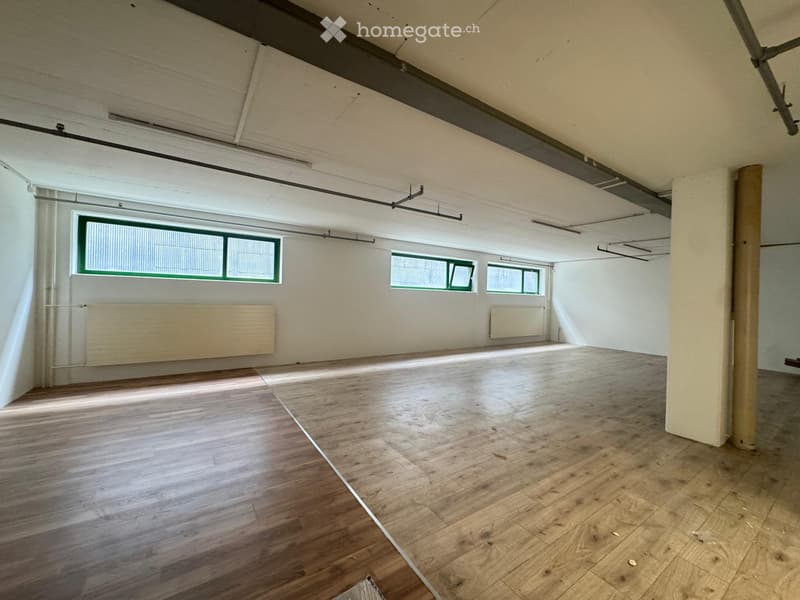 Empty room with wooden flooring, white walls, and large windows with green frames. Exposed ceiling beams and radiators are visible.