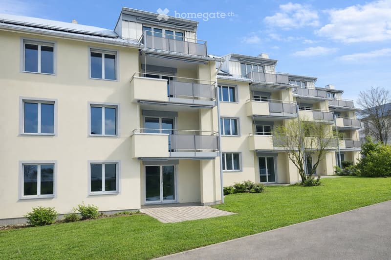 Modern multi-story residential building with light yellow facade, large windows, and multiple balconies. The building is surrounded by a well-maintained lawn and a paved driveway.