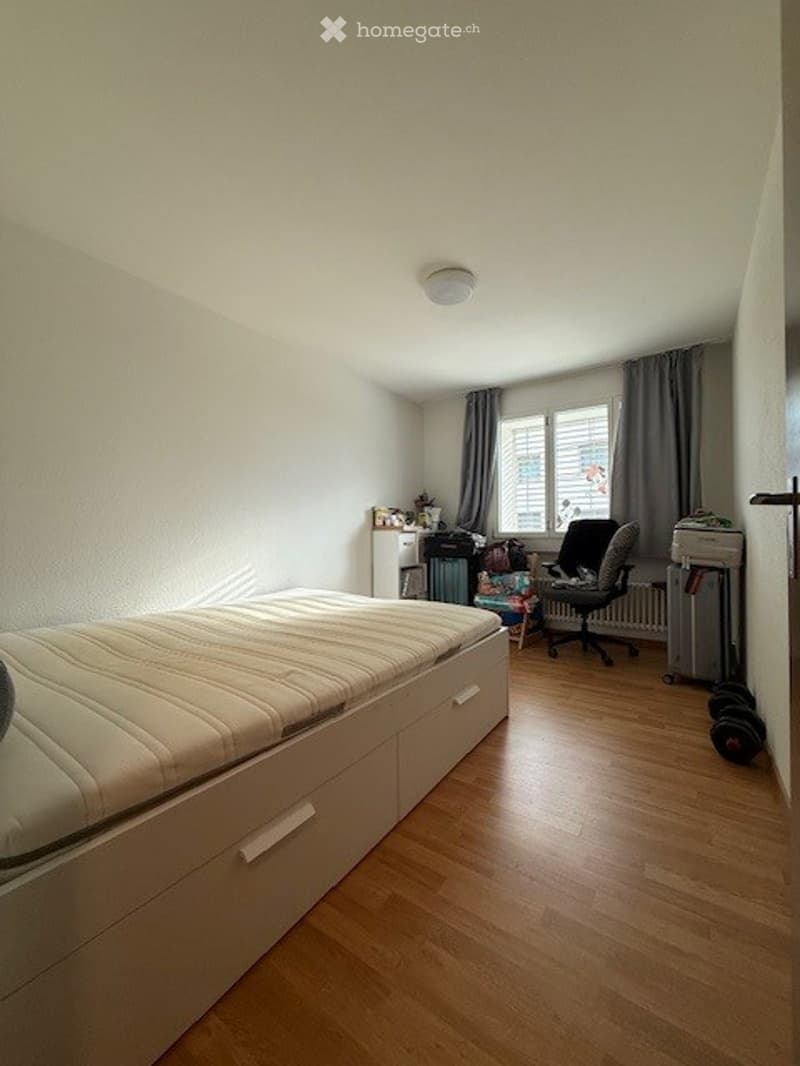 A tidy bedroom with a large bed, a chair, an office chair, a radiator, a storage unit, and luggage near the window.