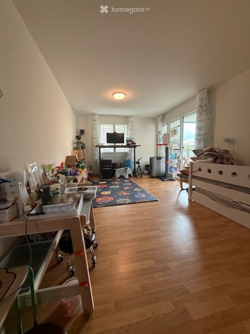 The room has wooden flooring, a sofa, a table, a chair, a screen, a laptop, a stool, and a fridge. There is also a scooter in the corner.