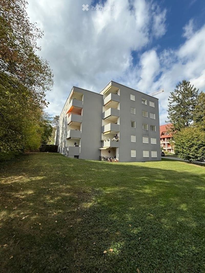 Modern multi-story apartment building with balconies, surrounded by green lawn and trees.