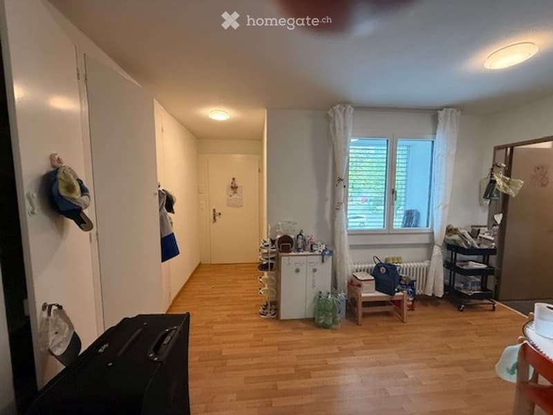 The room has wooden flooring, white walls, and a window with curtains. There is a small cabinet, a stool, a radiator, and a cart with items on it.