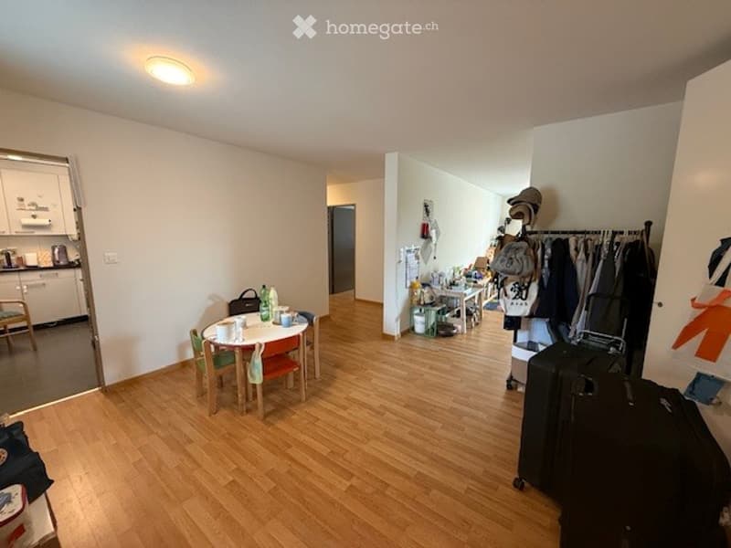 The image shows a furnished room with a dining table and chairs, a small kitchen area with a cupboard and stool, and a coat rack with bags and a suitcase.