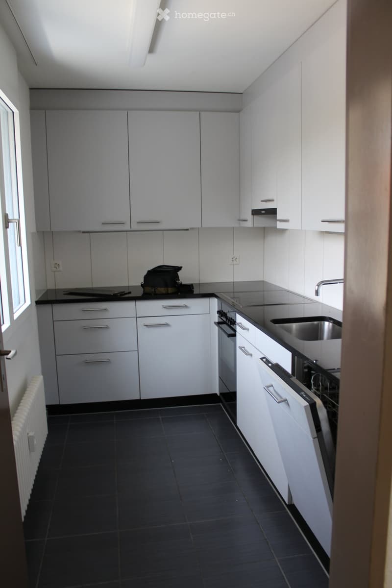 Modern kitchen with white cabinets, black countertops, stainless steel sink, and oven. Features include a window, radiator, and a backpack on the counter.