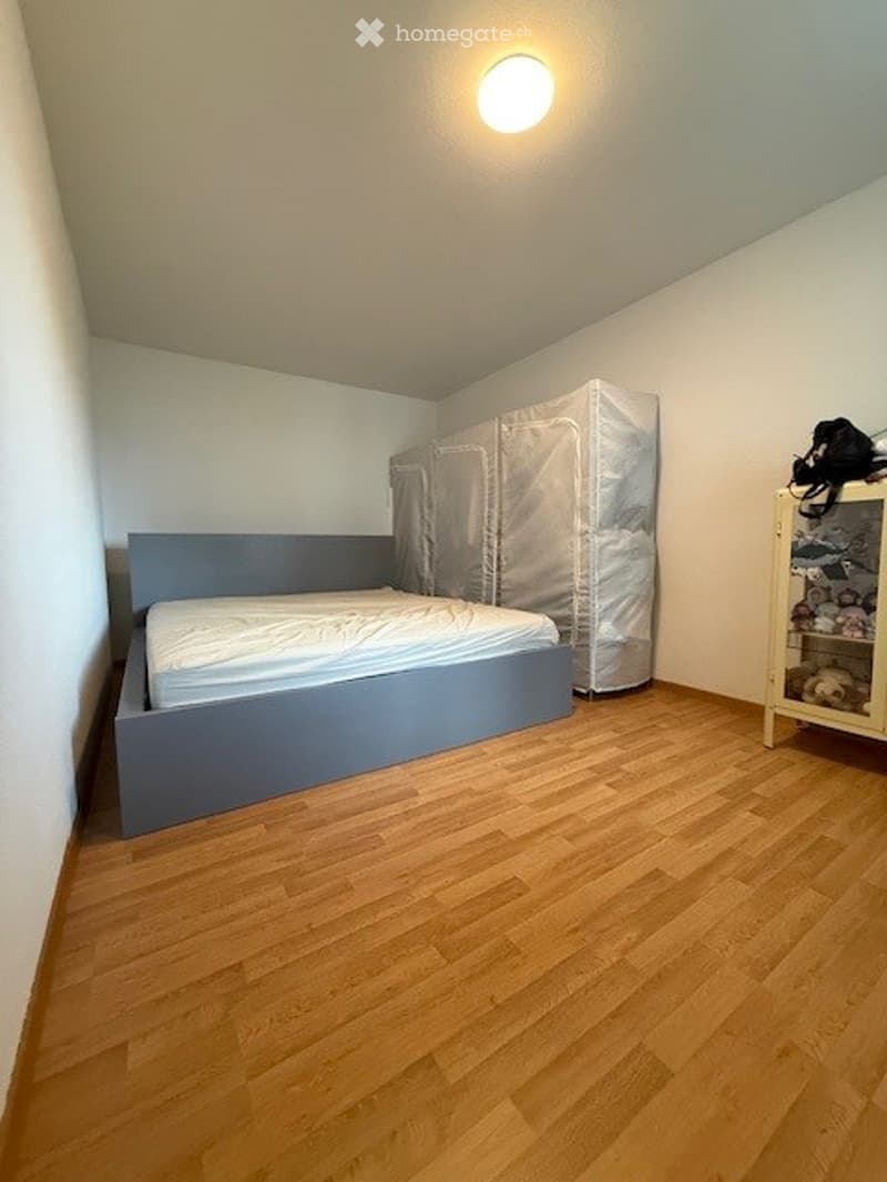 A small bedroom with a wooden floor, a grey bed frame with a white mattress, and a covered mattress on the floor. A small wardrobe with shelves is visible in the corner.