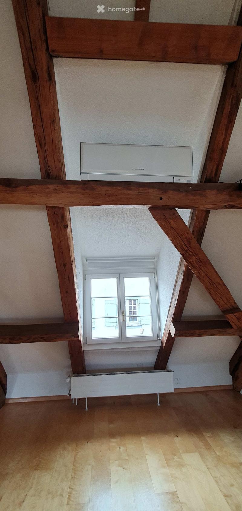 Empty room with wooden beams, wooden floor, white walls, and a window with a radiator below it.