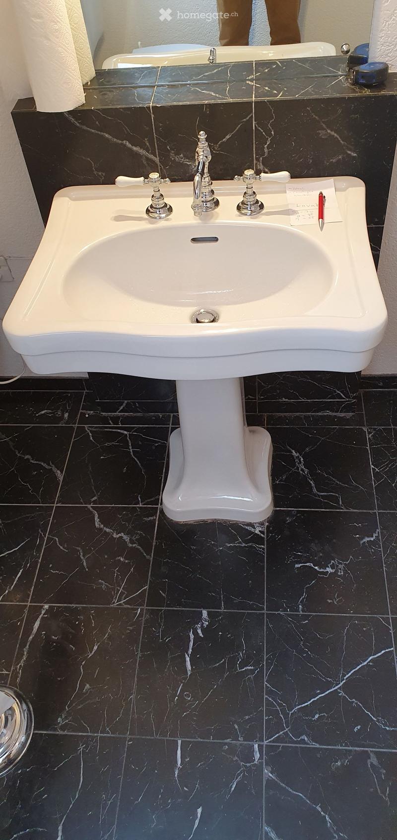 White pedestal sink with chrome faucet and handles, placed on dark marble tiles.
